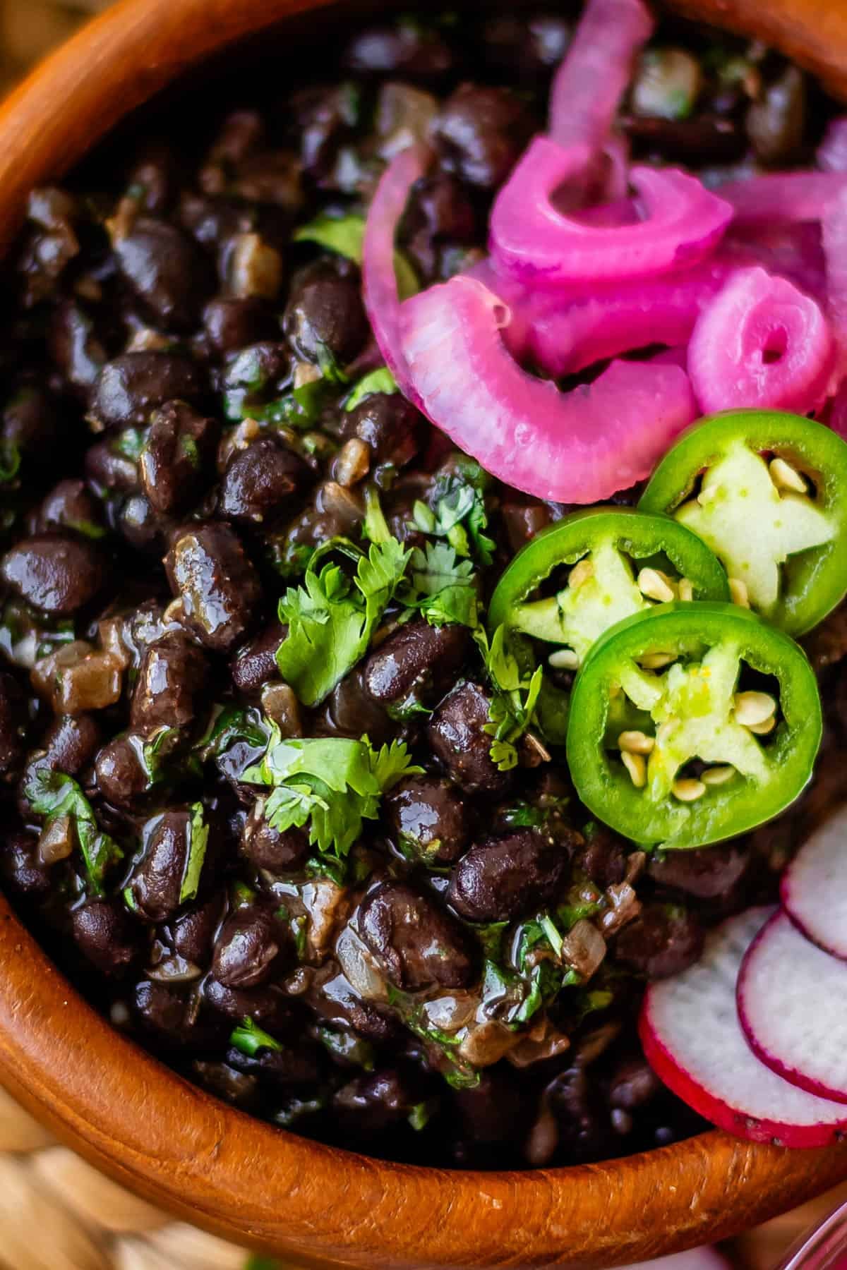 A bowl of black beans garnished with cilantro, sliced jalapeños, pickled red onions, and radish slices.