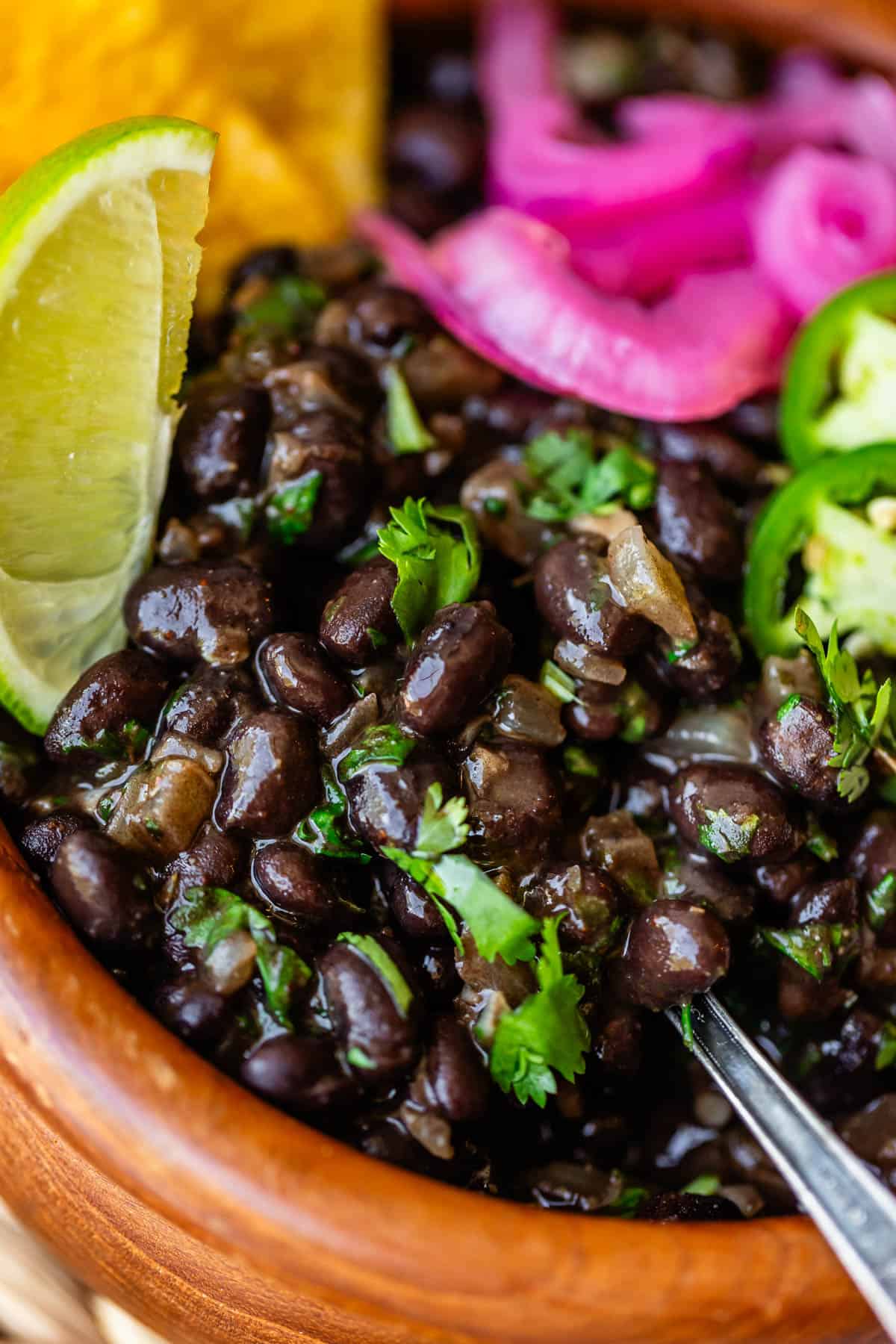 Close-up of a bowl of black beans garnished with fresh cilantro, lime wedges, sliced jalapeños, and pickled red onions, with a spoon in the bowl.