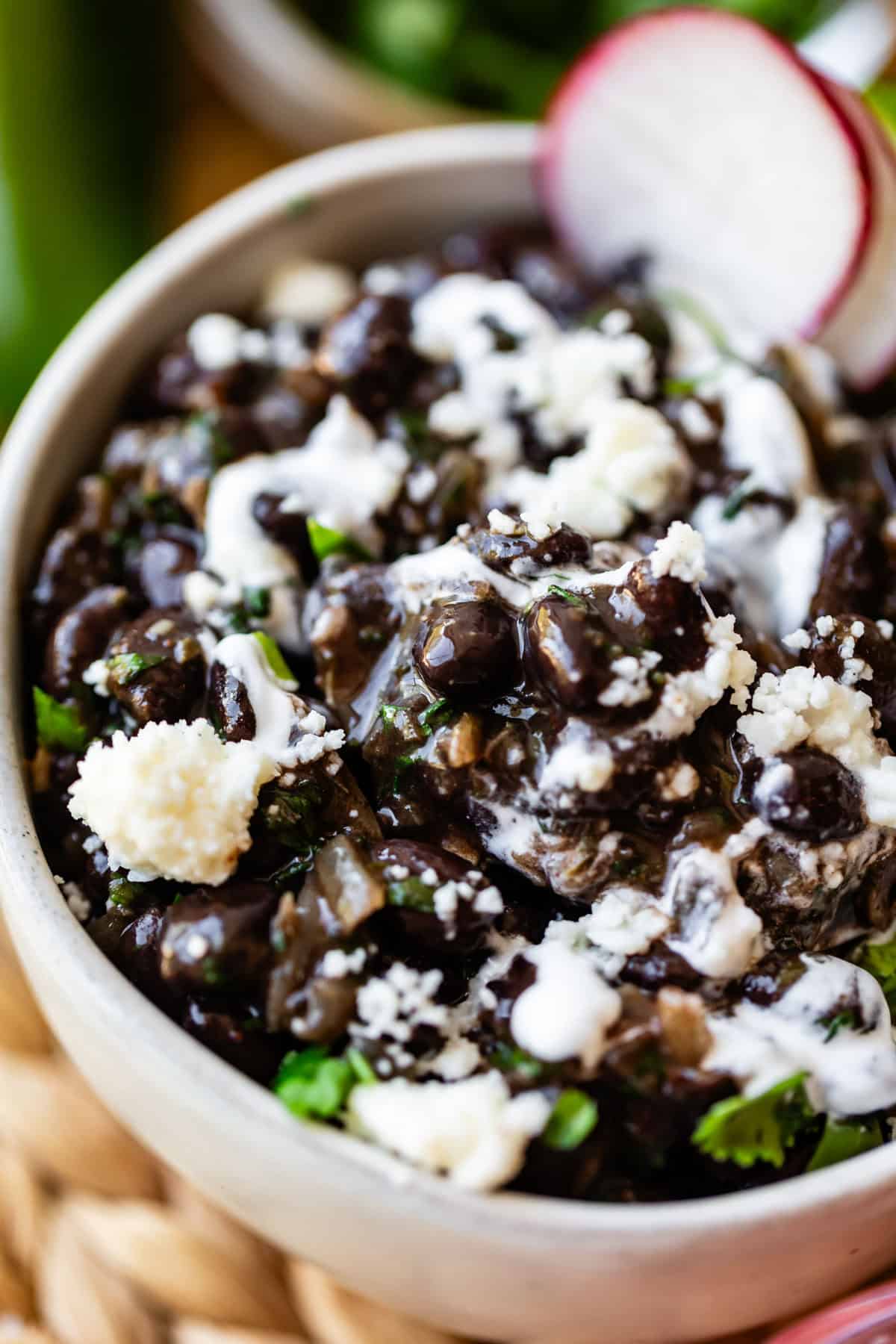 A close-up of a bowl filled with creamy black beans, garnished with crumbled cheese, fresh cilantro, and a drizzle of white sauce. A sliced radish sits on the edge of the bowl.