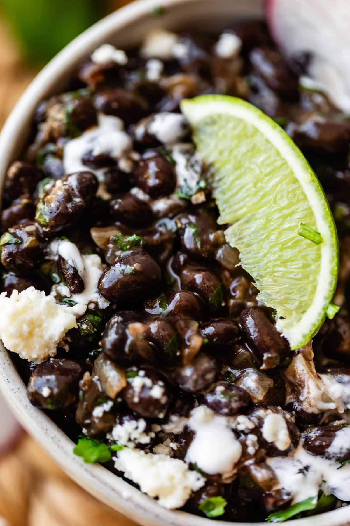 A close-up of a bowl filled with black beans, topped with crumbled cheese, a drizzle of white sauce, chopped herbs, and a wedge of lime.