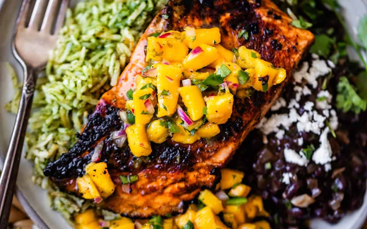 Grilled salmon topped with mango salsa, served with green rice and black beans, garnished with cheese and herbs; a fork rests on the plates edge.