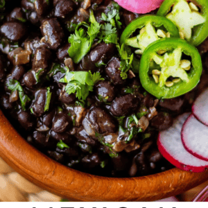 A bowl of Mexican black beans garnished with cilantro, sliced jalapeños, pickled onions, and radish slices, with text reading Mexican Black Beans The Food Charlatan..