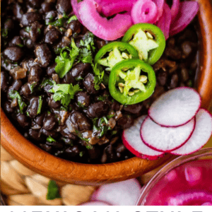 A bowl of Mexican-style black beans topped with pickled onions, sliced jalapeños, and cilantro, with sliced radishes and herbs on the side.