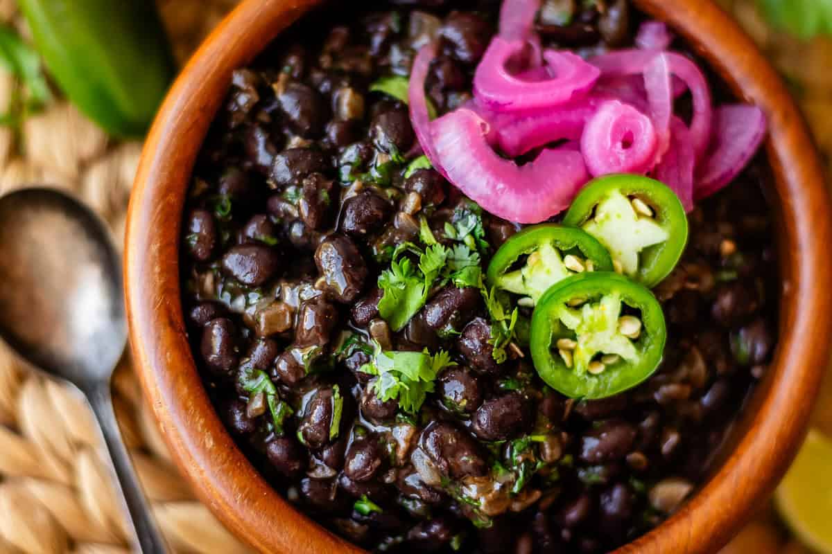 A bowl of black beans garnished with fresh cilantro, sliced jalapeños, and pickled red onions, with a spoon beside the bowl on a woven surface.