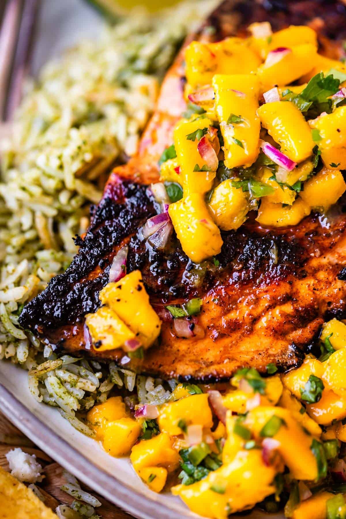 Grilled chicken topped with mango salsa, served with green herb rice. The salsa includes diced mango, red onion, and herbs, adding color and freshness to the dish.