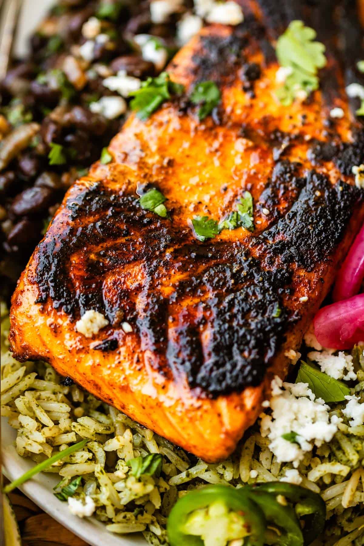 Grilled salmon fillet with char marks sits atop seasoned rice, garnished with cilantro, sliced jalapeños, black beans, and crumbled cheese.