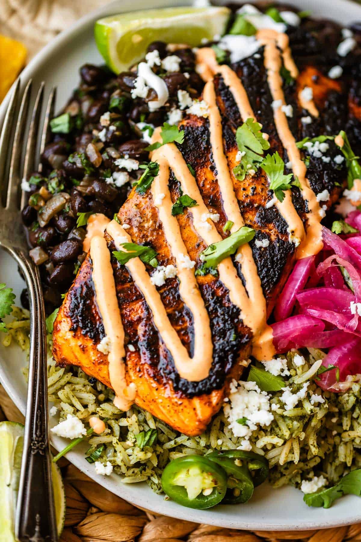 A grilled salmon fillet with sauce sits on green rice, next to black beans, pickled onions, jalapeños, cilantro, and lime wedges on a plate, with a fork on the side.