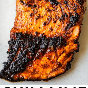 A close-up of a grilled, blackened salmon fillet with a chili lime crust, charred edges, and a vibrant orange color on a white background. Text below reads: Chili Lime Blackened Salmon. The Food Charlatan..