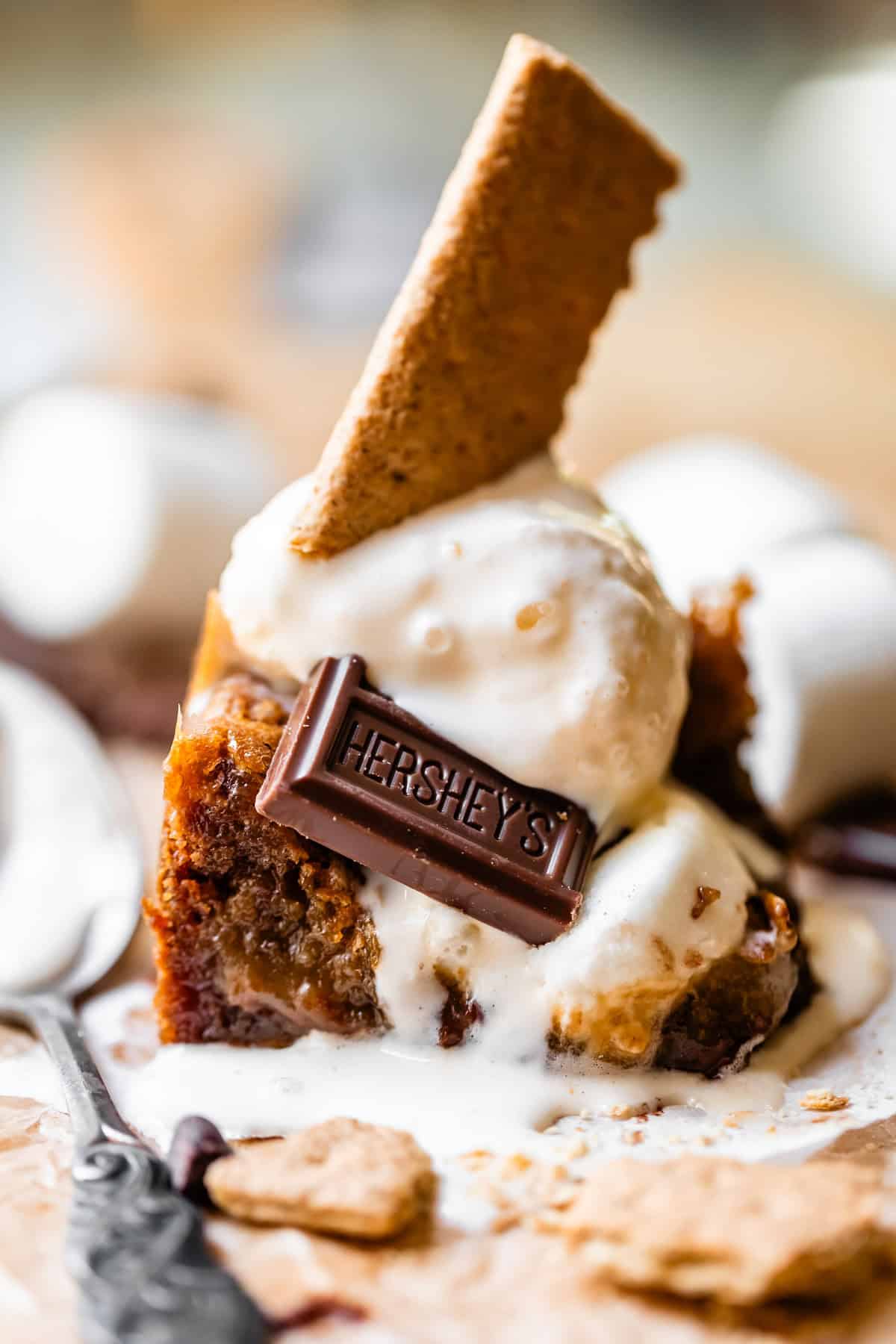 A dessert with melting vanilla ice cream, a Hersheys chocolate piece, and a graham cracker on top of a gooey brownie, with marshmallows in the background.