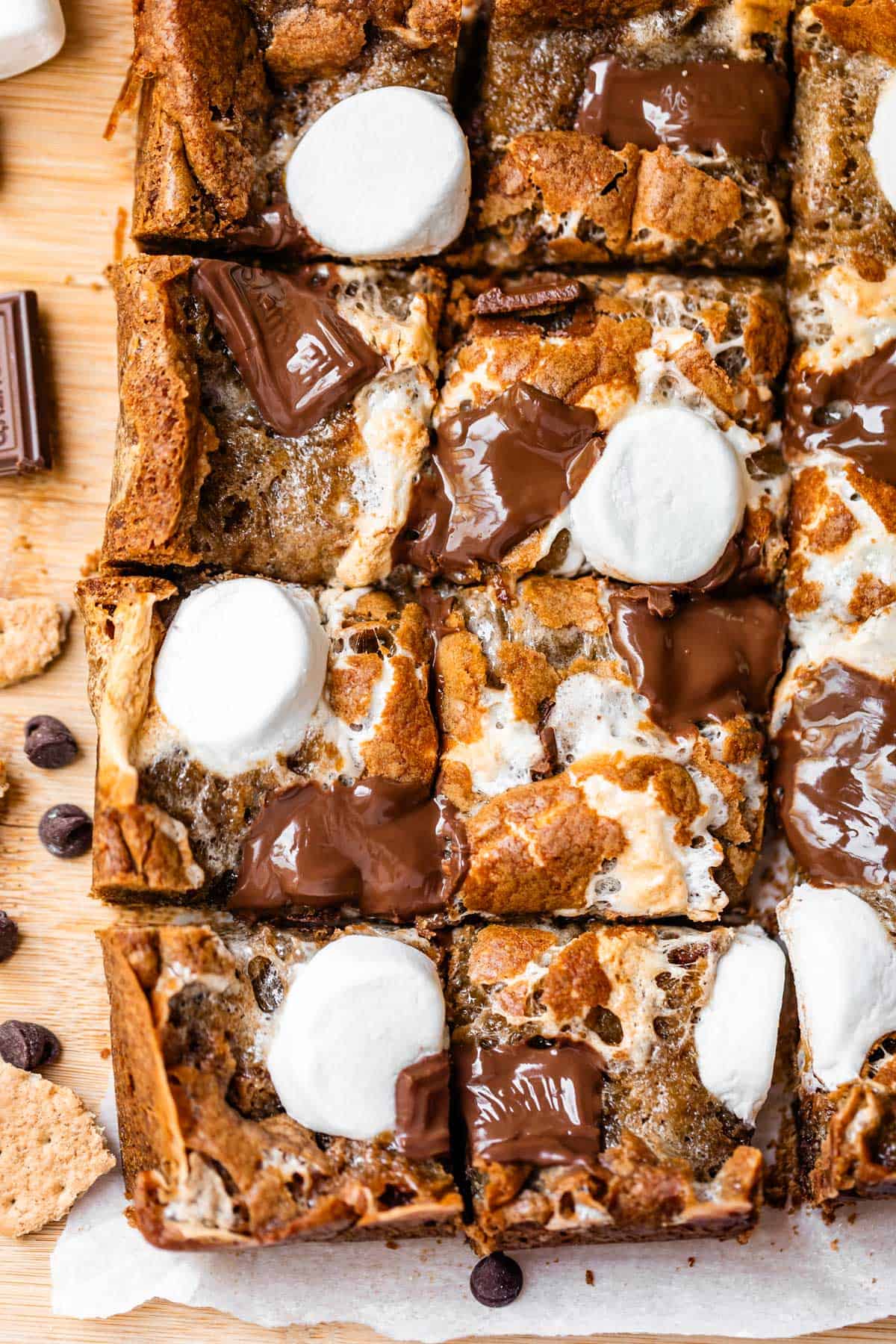 A close-up of smores bars cut into squares, topped with melted chocolate pieces and toasted marshmallows on a wooden surface.