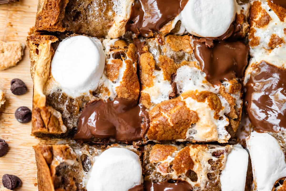 Close-up of gooey smores bars topped with melted chocolate chunks and toasted marshmallows, cut into squares on a wooden surface with scattered chocolate chips nearby.
