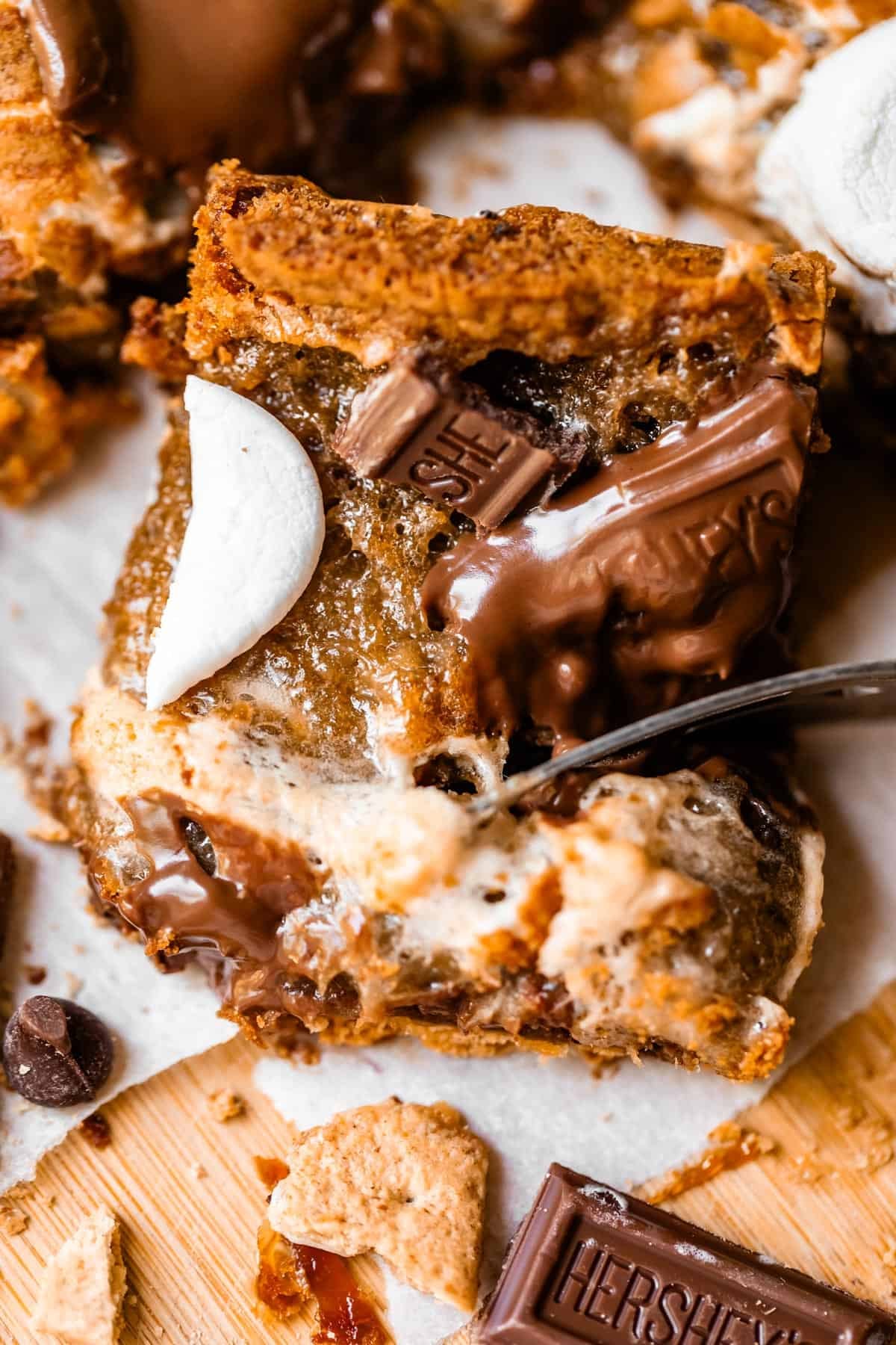 Close-up of a gooey smores bar topped with melted chocolate, graham cracker pieces, and toasted marshmallow, sitting on parchment paper with bits of chocolate and crumbs around it.