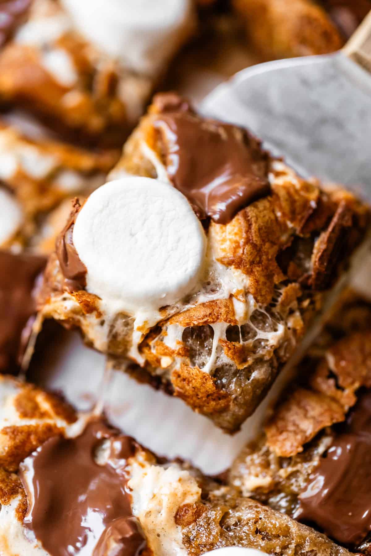 A close-up of a gooey smores bar topped with melted chocolate and a toasted marshmallow, being lifted by a spatula.