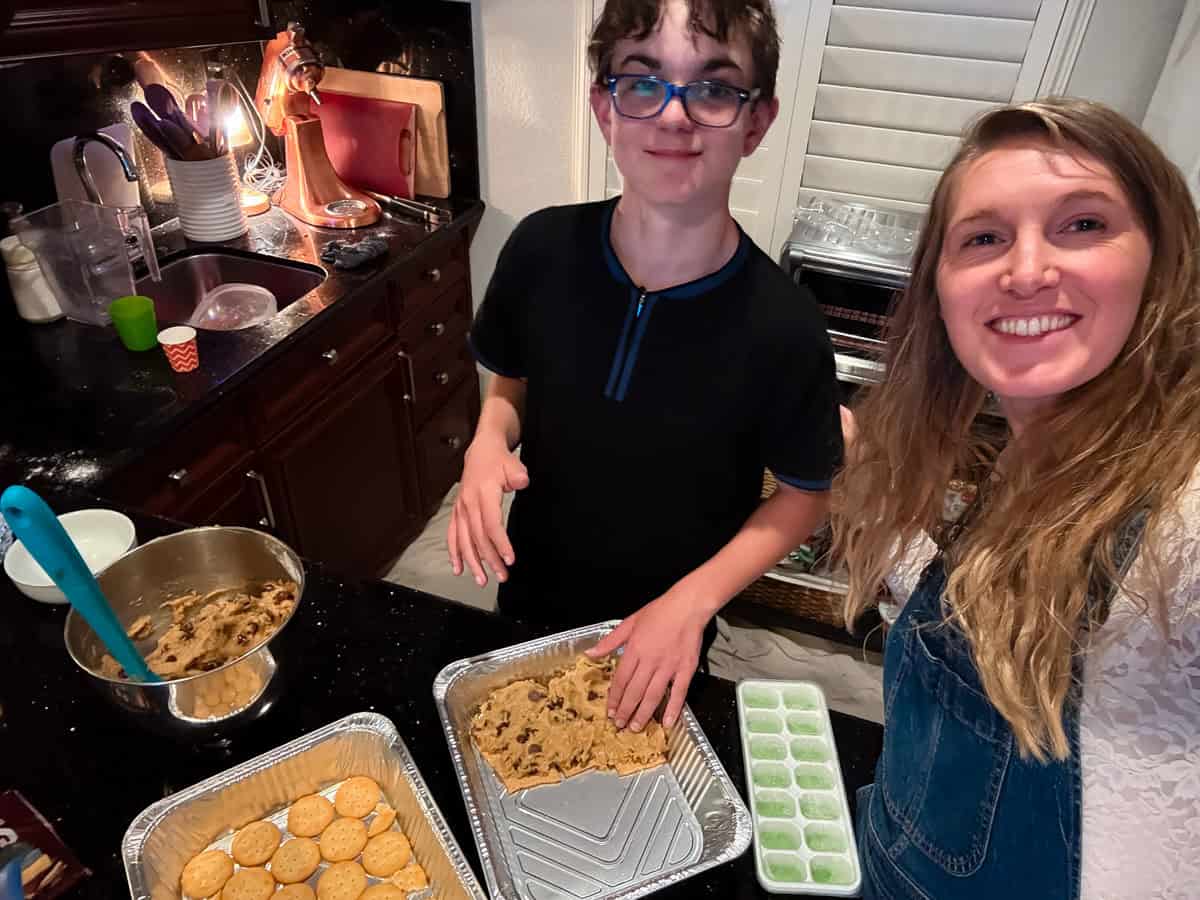 A woman and a boy smile while preparing cookie dough and graham crackers in a kitchen, with baking trays, bowls, and utensils on the counter.
