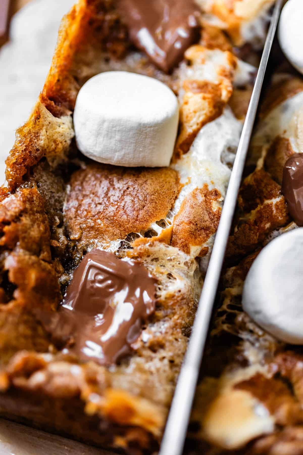 Close-up of a baked dessert with golden brown crust, topped with melted chocolate squares and toasted marshmallows, being sliced with a knife.