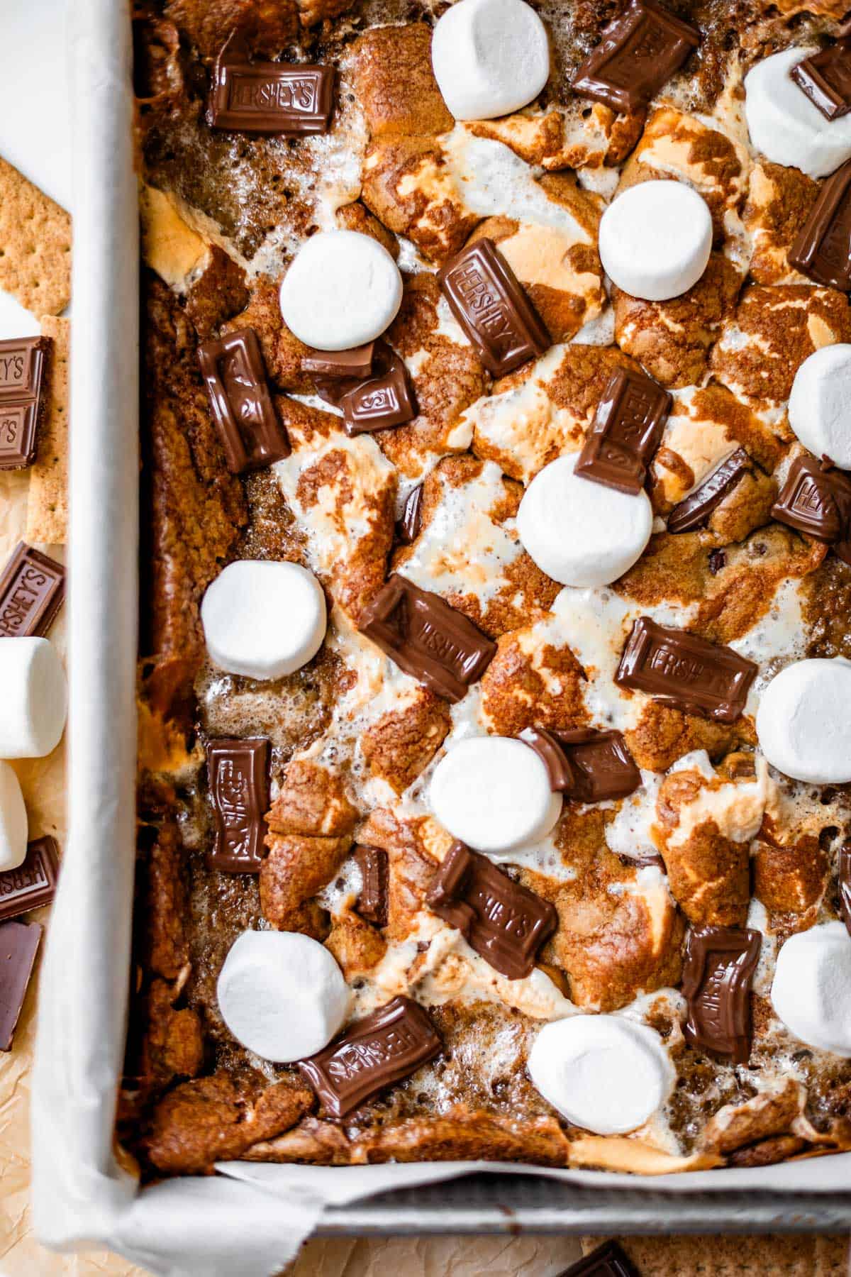 A baked smores dessert bar in a pan, topped with toasted marshmallows and pieces of chocolate, next to graham crackers and extra marshmallows on the side.