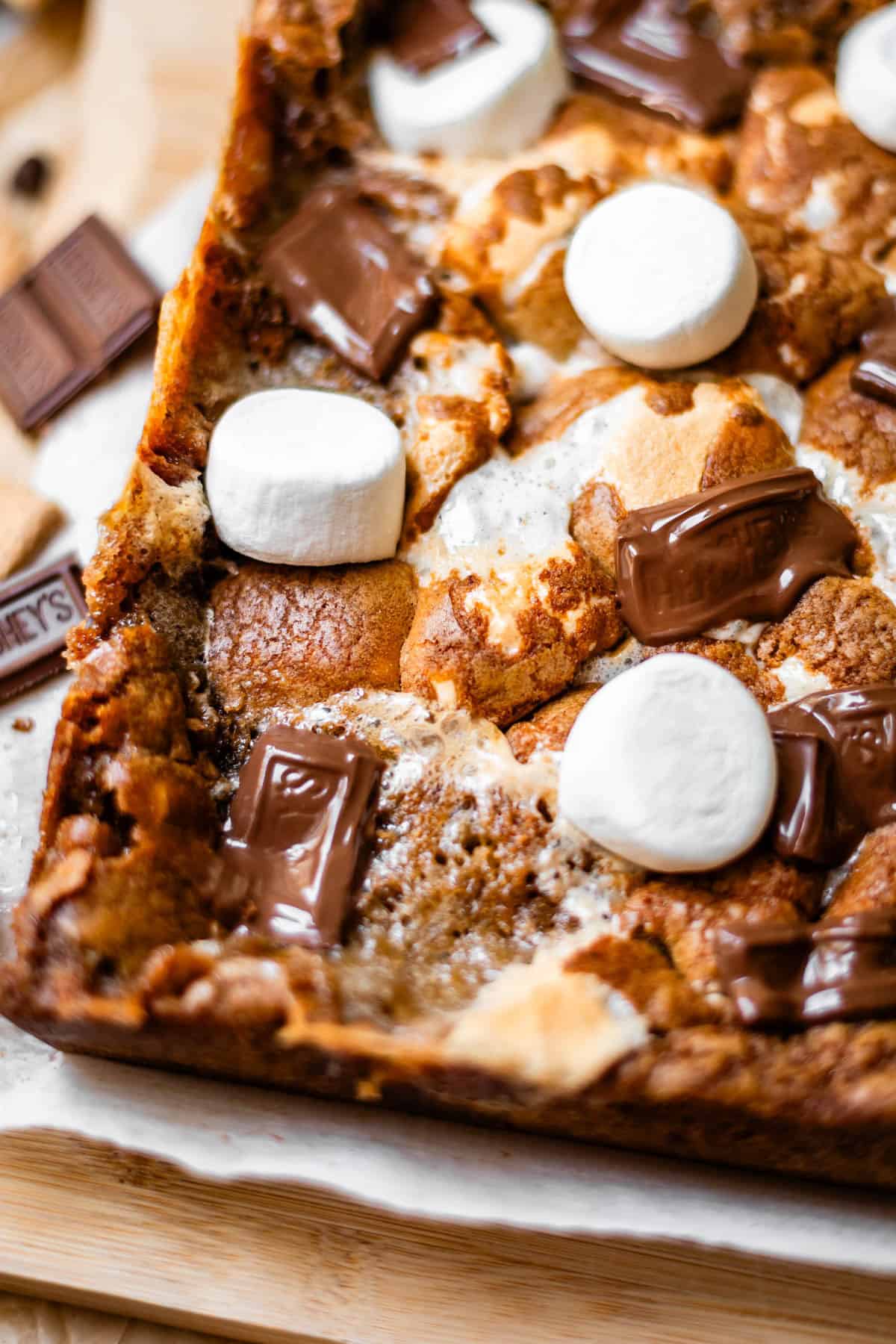 A close-up of baked smores bars topped with toasted marshmallows and melted chocolate pieces on a wooden surface.