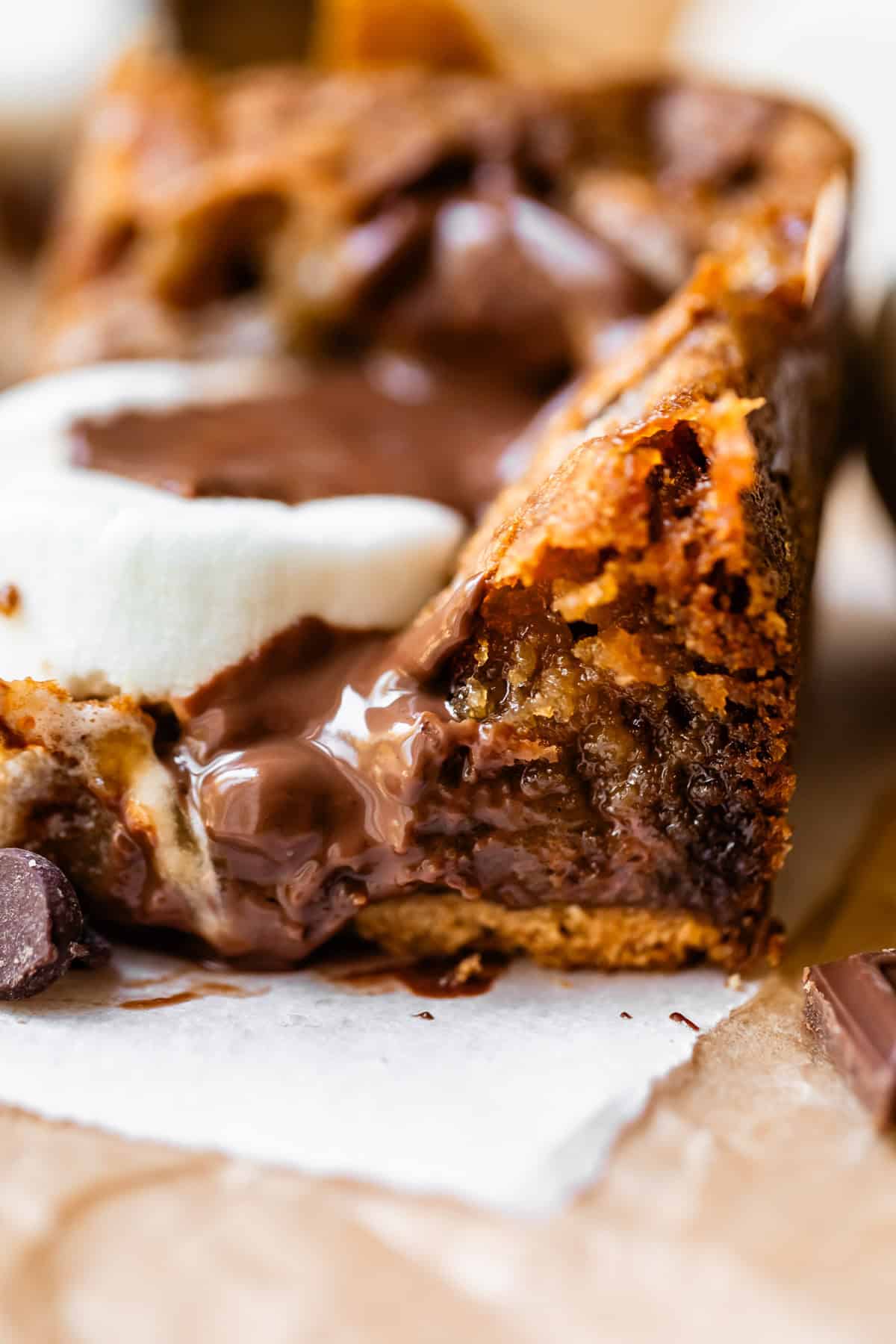 A close-up of a gooey chocolate chip cookie bar topped with melted chocolate and a slice of marshmallow, resting on parchment paper.