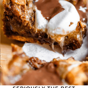 Close-up of gooey smores cookie bars with melted chocolate and toasted marshmallow, set on parchment paper. Text below reads: Seriously the Best Smores Cookie Bars - The Food Charlatan..