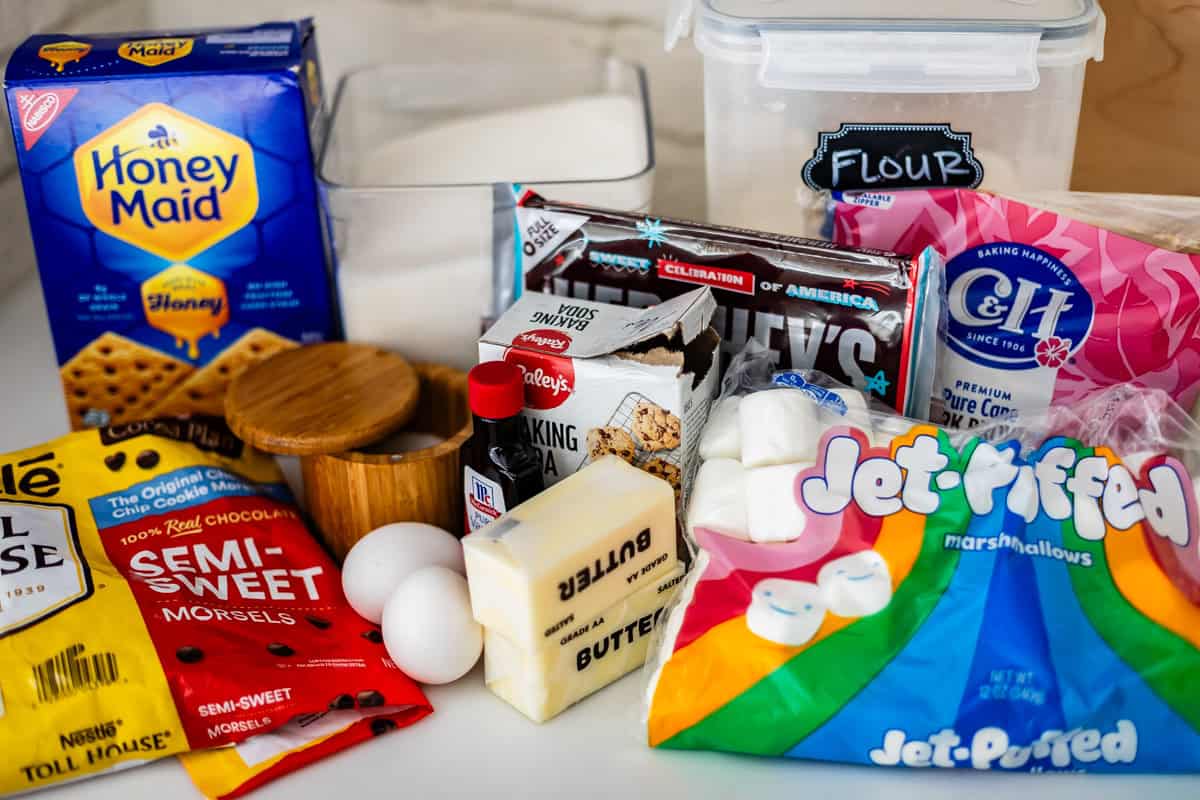 A variety of baking ingredients on a countertop, including graham crackers, sugar, flour, eggs, butter, chocolate chips, marshmallows, baking powder, vanilla, and salt.
