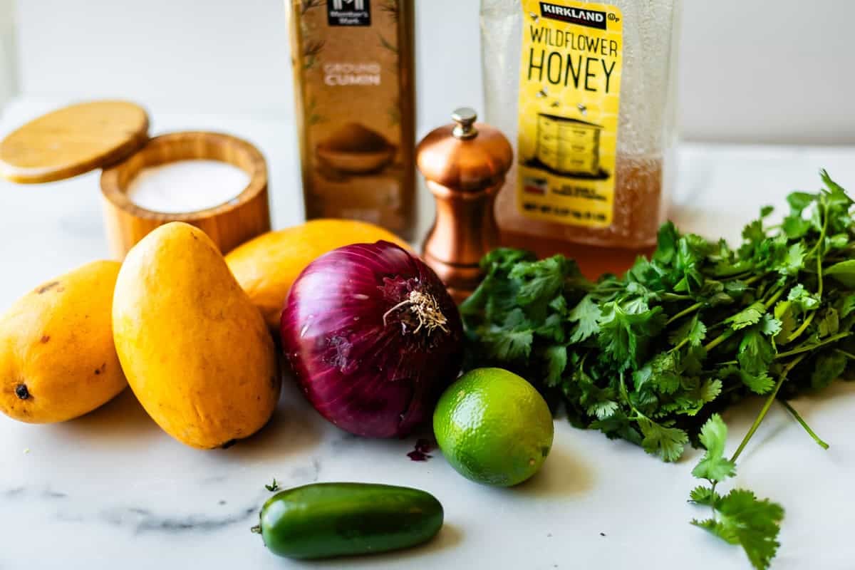 Three mangoes, a red onion, cilantro, a lime, a jalapeño, a salt container, ground cumin, pepper grinder, and a bottle of wildflower honey on a white surface.