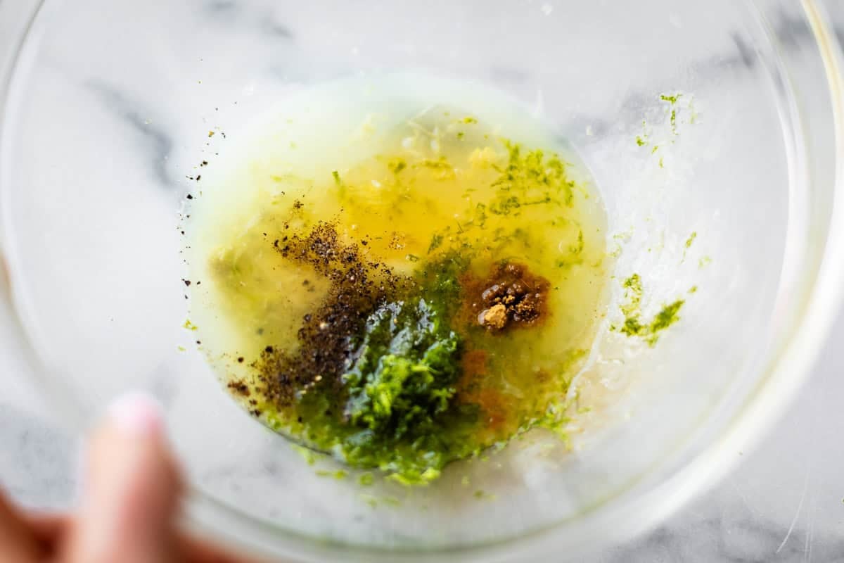 A glass bowl with oil, lime zest, ground pepper, a brown spice, and a green herb mixture on a marble surface, ready to be mixed.