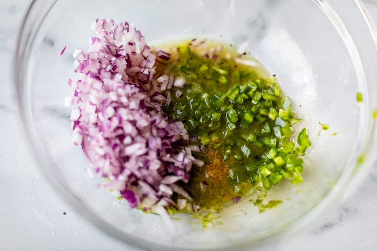 A glass bowl with finely chopped red onion, chopped green jalapeño, and a pool of greenish liquid on a white surface.