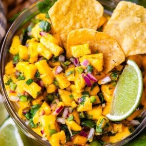 A bowl of fresh mango salsa with diced mango, red onion, cilantro, and jalapeño, garnished with lime wedges and served with tortilla chips.