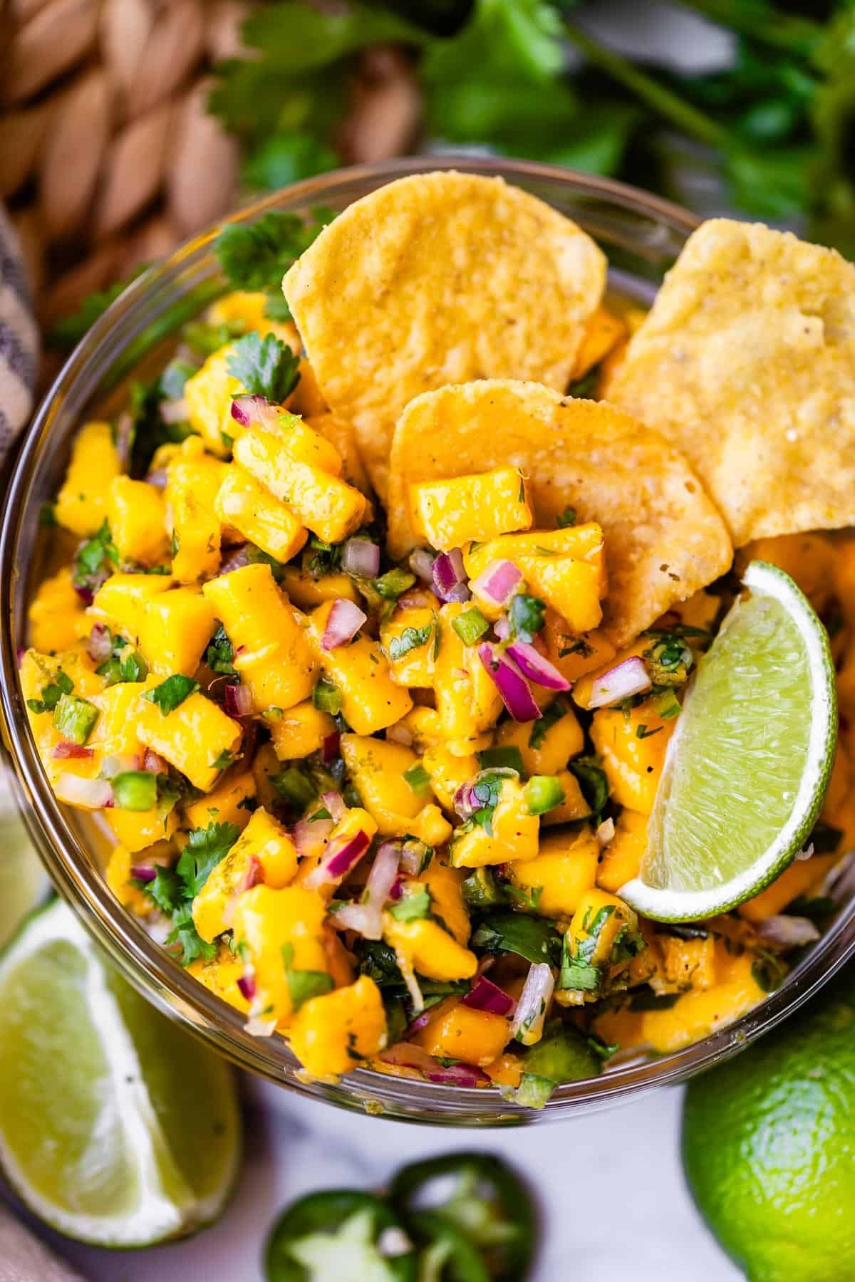 A bowl of fresh mango salsa with diced mango, red onion, cilantro, and jalape&ntilde;o, garnished with lime wedges and served with tortilla chips.