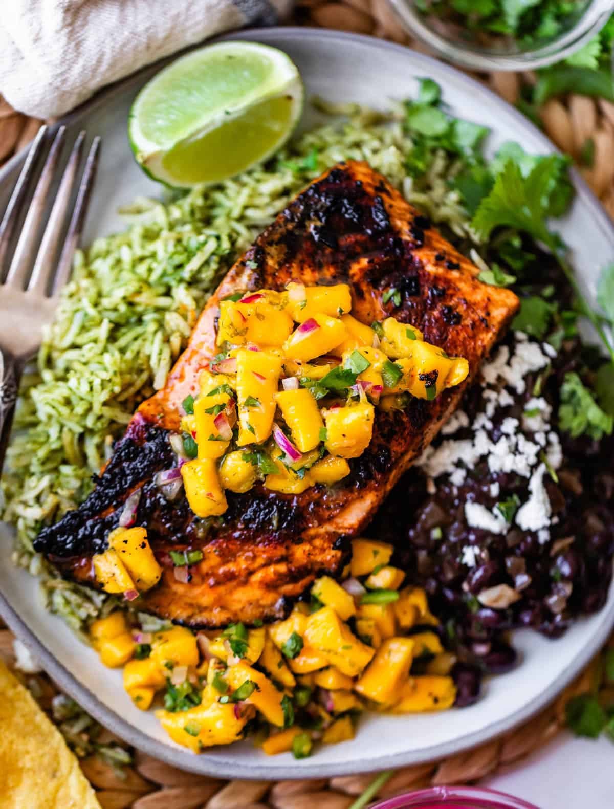 A plate with grilled salmon topped with mango salsa, served over green rice, with black beans, crumbled cheese, cilantro, and a lime wedge.