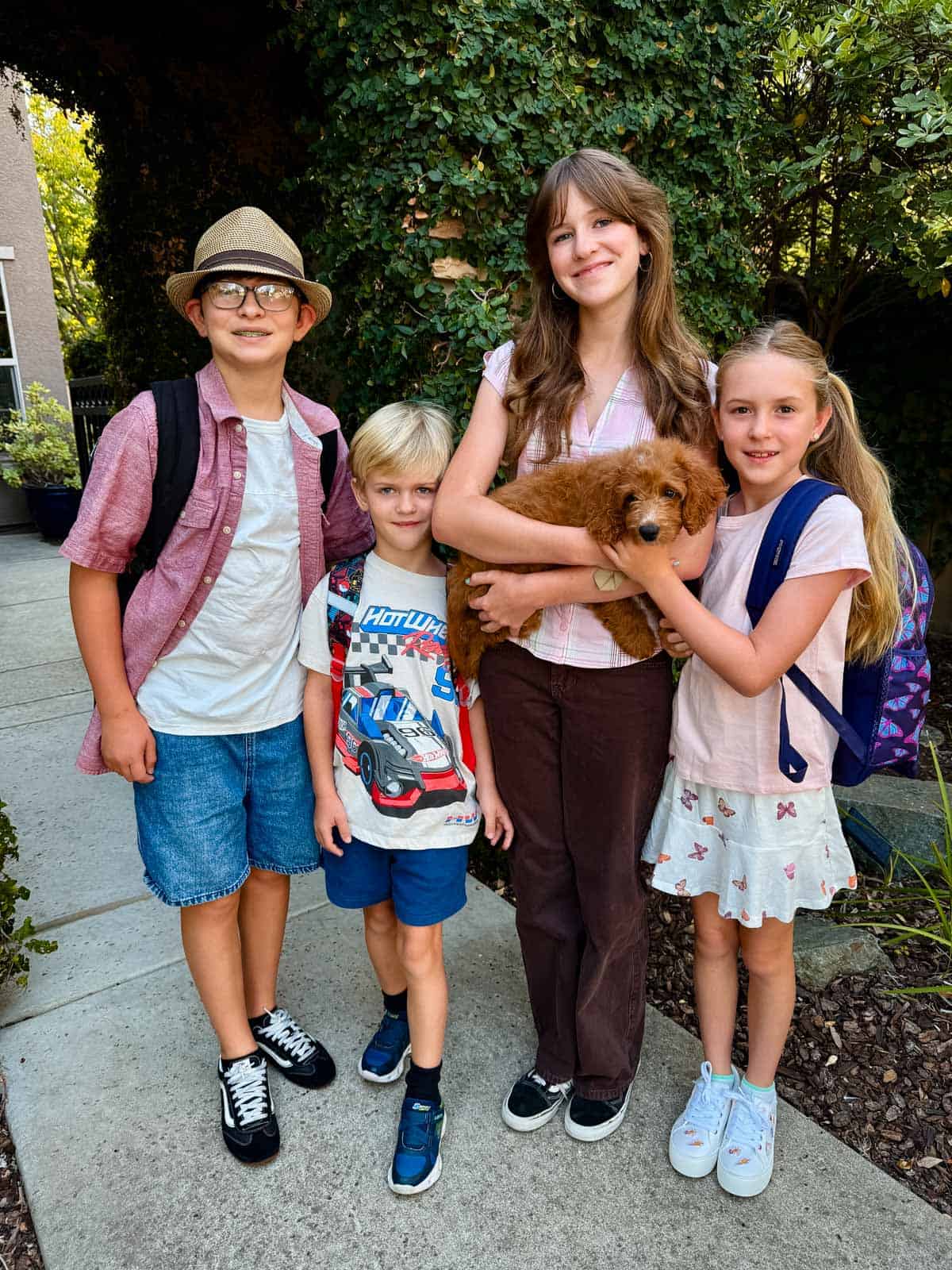 Four children stand outdoors with backpacks; one girl in the center is holding a small brown puppy. They are smiling and surrounded by greenery.