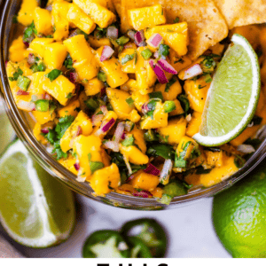 A bowl of mango salsa with tortilla chips and a lime wedge, surrounded by fresh cilantro and limes. Text reads: This mango salsa is legit. The Food Charlatan.