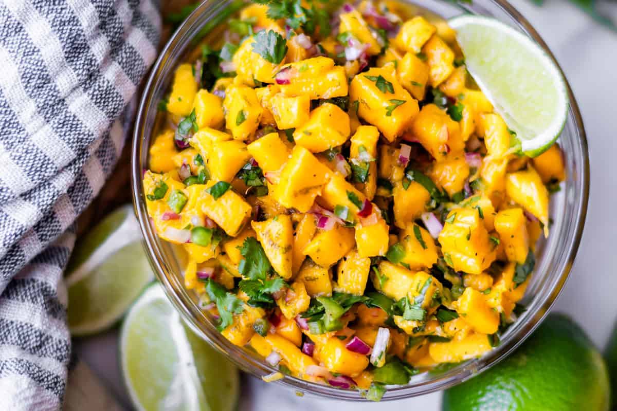A glass bowl filled with vibrant mango salsa made of diced mango, red onion, cilantro, and lime wedges, sits next to a striped towel and more lime slices.
