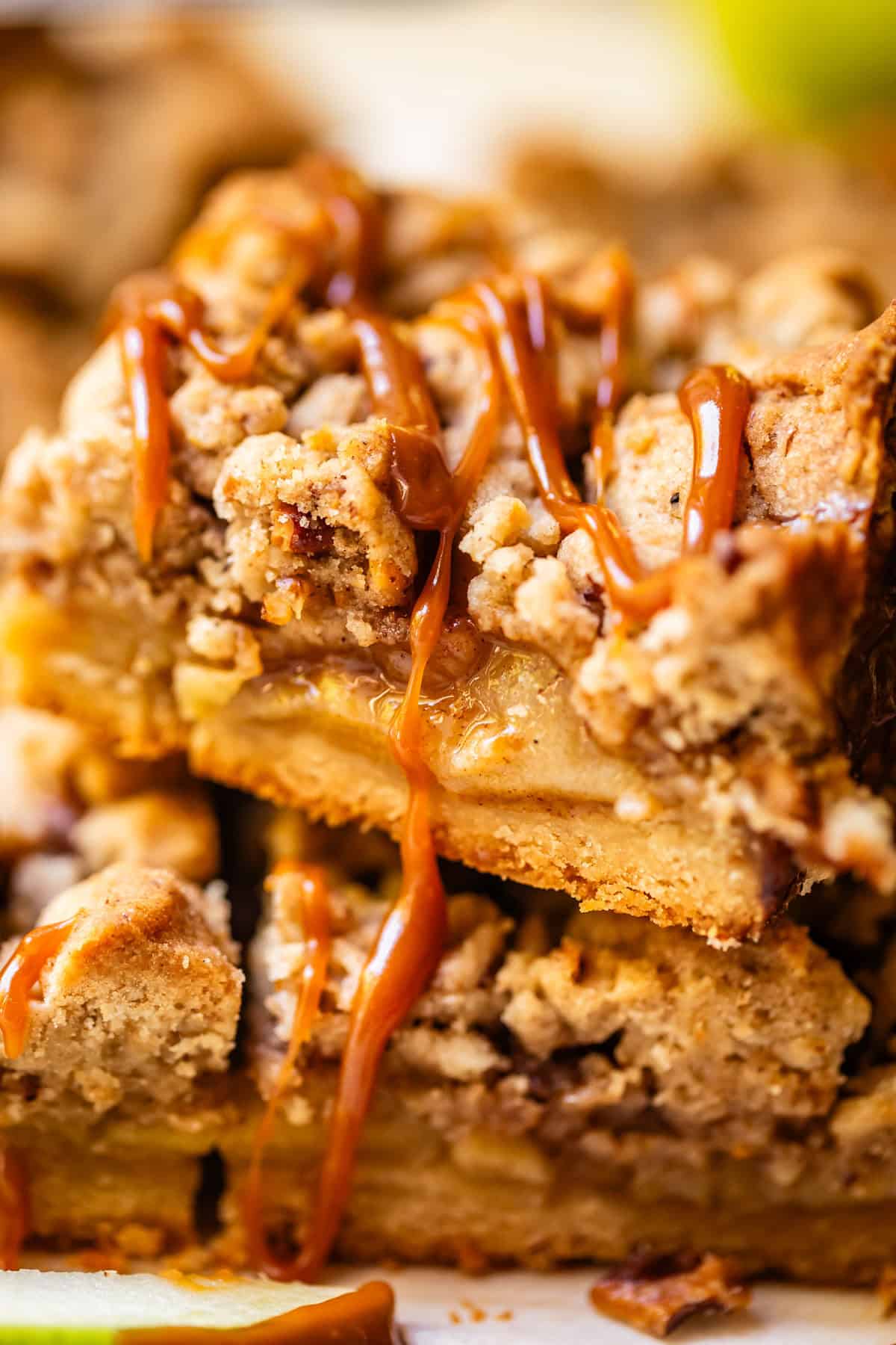 Close-up of crumbly dessert bars with a layer of apples and crumb topping, drizzled with caramel sauce. The bars are stacked, showing their moist interior and textured topping.