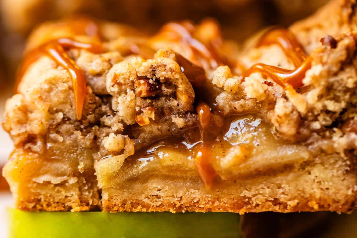 A close-up of a dessert bar with crumbly topping, gooey caramel drizzle, and layers of apple filling on a thick crust, sitting on a slice of green apple.