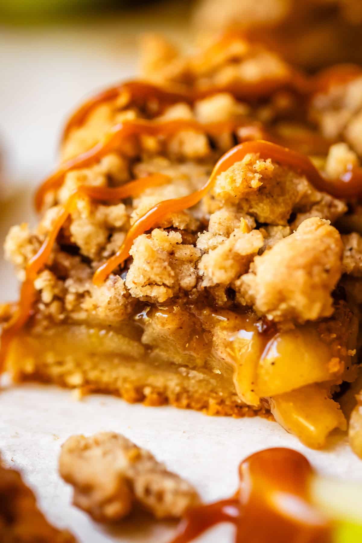 A close-up of a slice of apple crumble pie with a golden crumb topping, drizzled with caramel sauce, showing layers of cooked apples and crumbly crust.