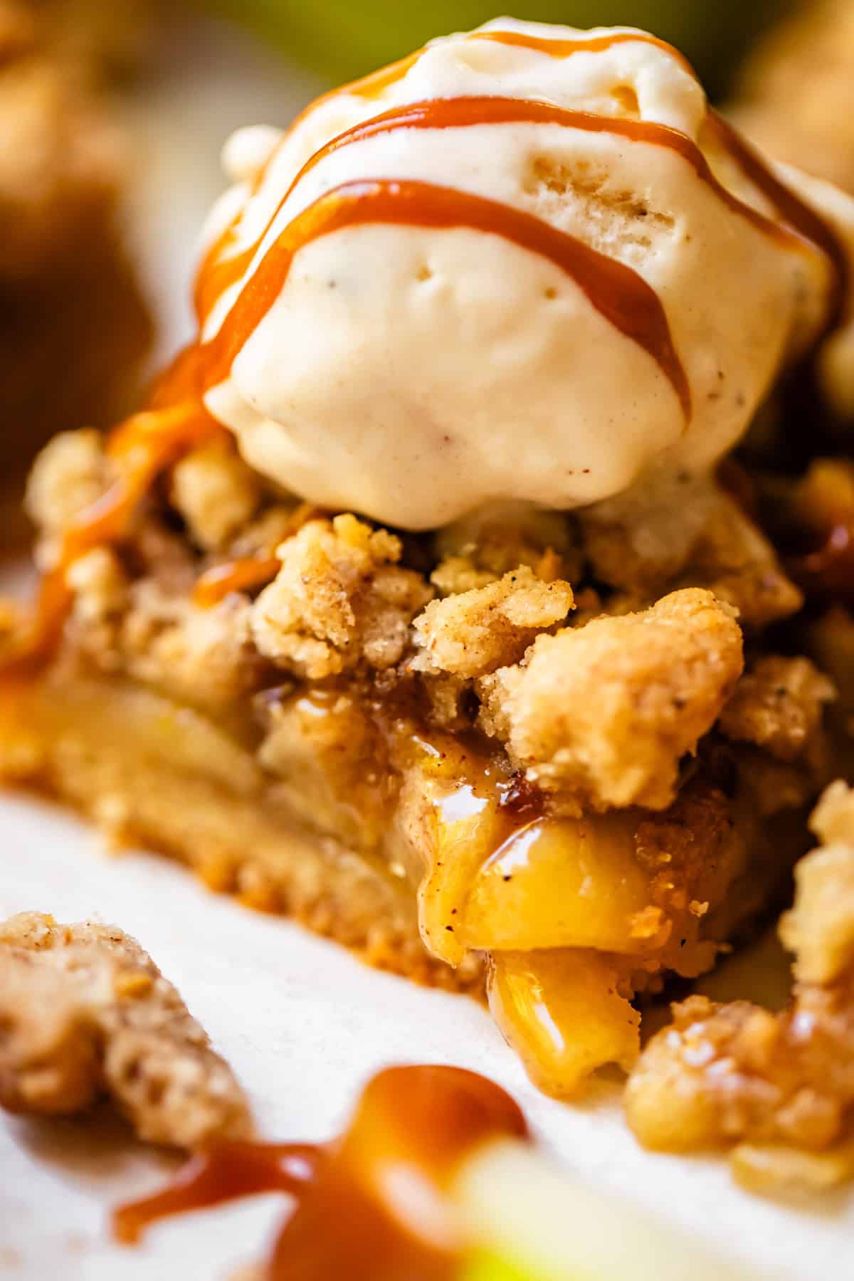 A close-up of apple crumble pie topped with a scoop of vanilla ice cream and drizzled with caramel sauce.