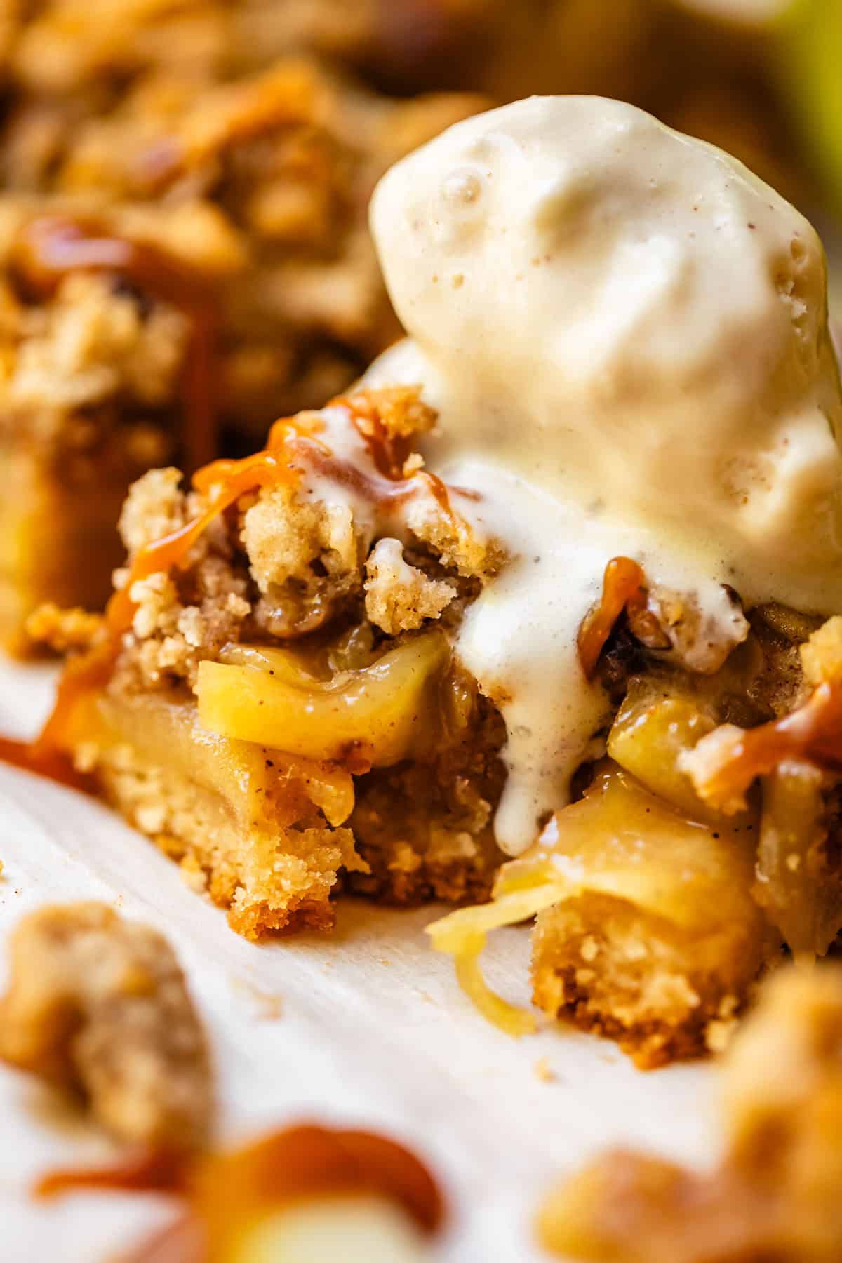 Close-up of a slice of apple crumble topped with melting vanilla ice cream and drizzled with caramel sauce. The dessert shows chunks of apple, crumbly topping, and a golden, buttery crust.
