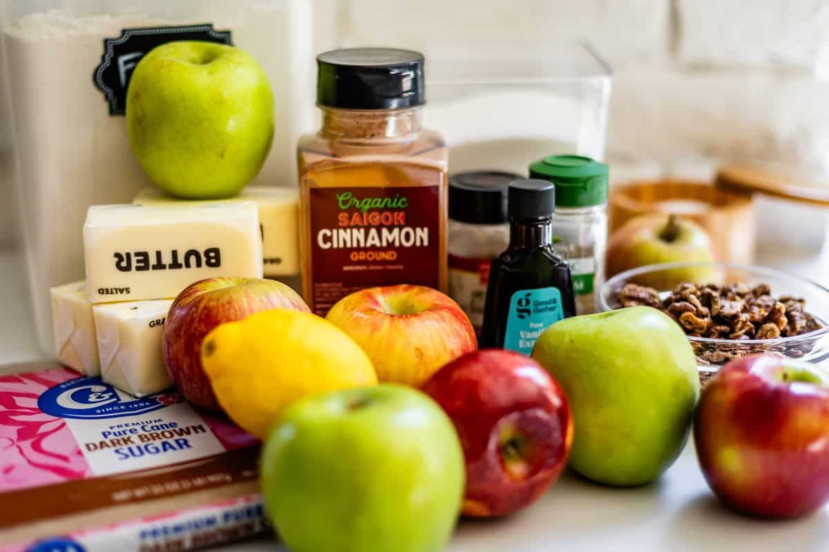 Apples, a lemon, butter, dark brown sugar, ground cinnamon, vanilla extract, nuts, and other baking ingredients are arranged on a kitchen counter.