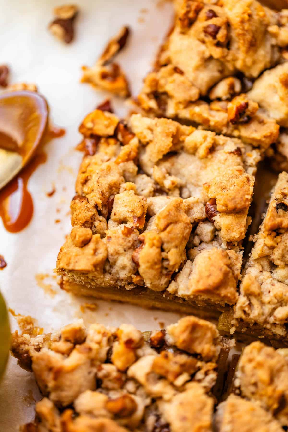 A close-up of crumbly dessert bars with chunks of nuts on parchment paper, next to a spoon with caramel sauce.