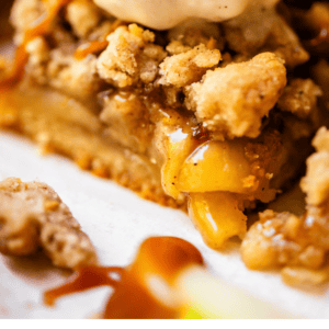 Close-up of apple pie bars topped with crumb topping, a scoop of vanilla ice cream, and caramel drizzle. Text below reads Perfect Holiday Apple Pie Bars – The Food Charlatan..