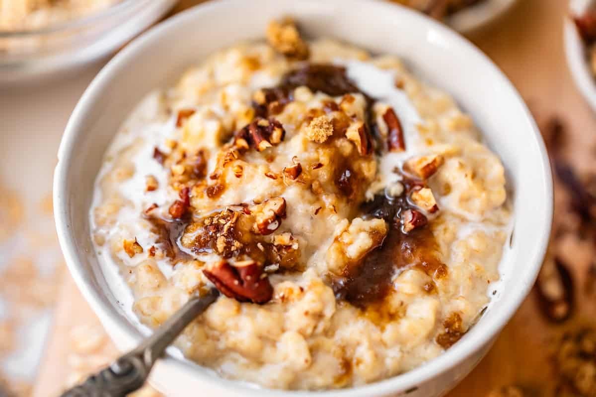 A bowl of oatmeal topped with chopped pecans, a drizzle of syrup, and a splash of milk, with a spoon resting inside.