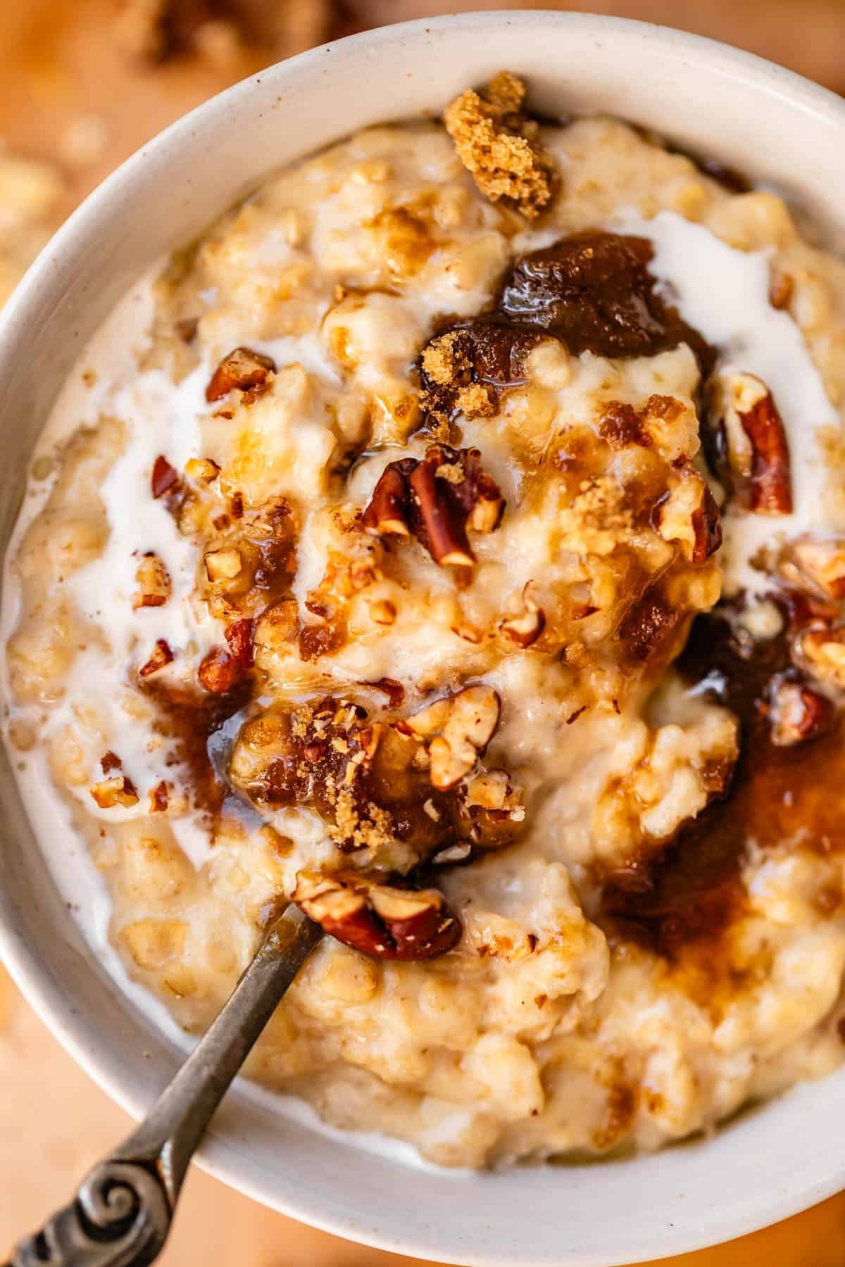 A bowl of oatmeal topped with brown sugar, pecans, and a swirl of cream, with a spoon resting inside.