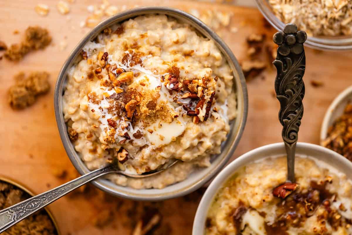 A bowl of oatmeal topped with brown sugar, chopped nuts, cream, and a pat of melting butter, with a spoon resting inside. Another similar bowl is nearby on a wooden surface.