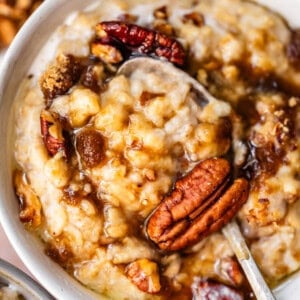 A close-up of a bowl of oatmeal topped with pecans, brown sugar, and syrup. A spoon is partially dipped in the creamy oatmeal, showcasing the rich toppings.