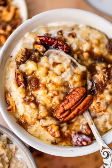 A close-up of a bowl of oatmeal topped with pecans, brown sugar, and syrup. A spoon is partially dipped in the creamy oatmeal, showcasing the rich toppings.