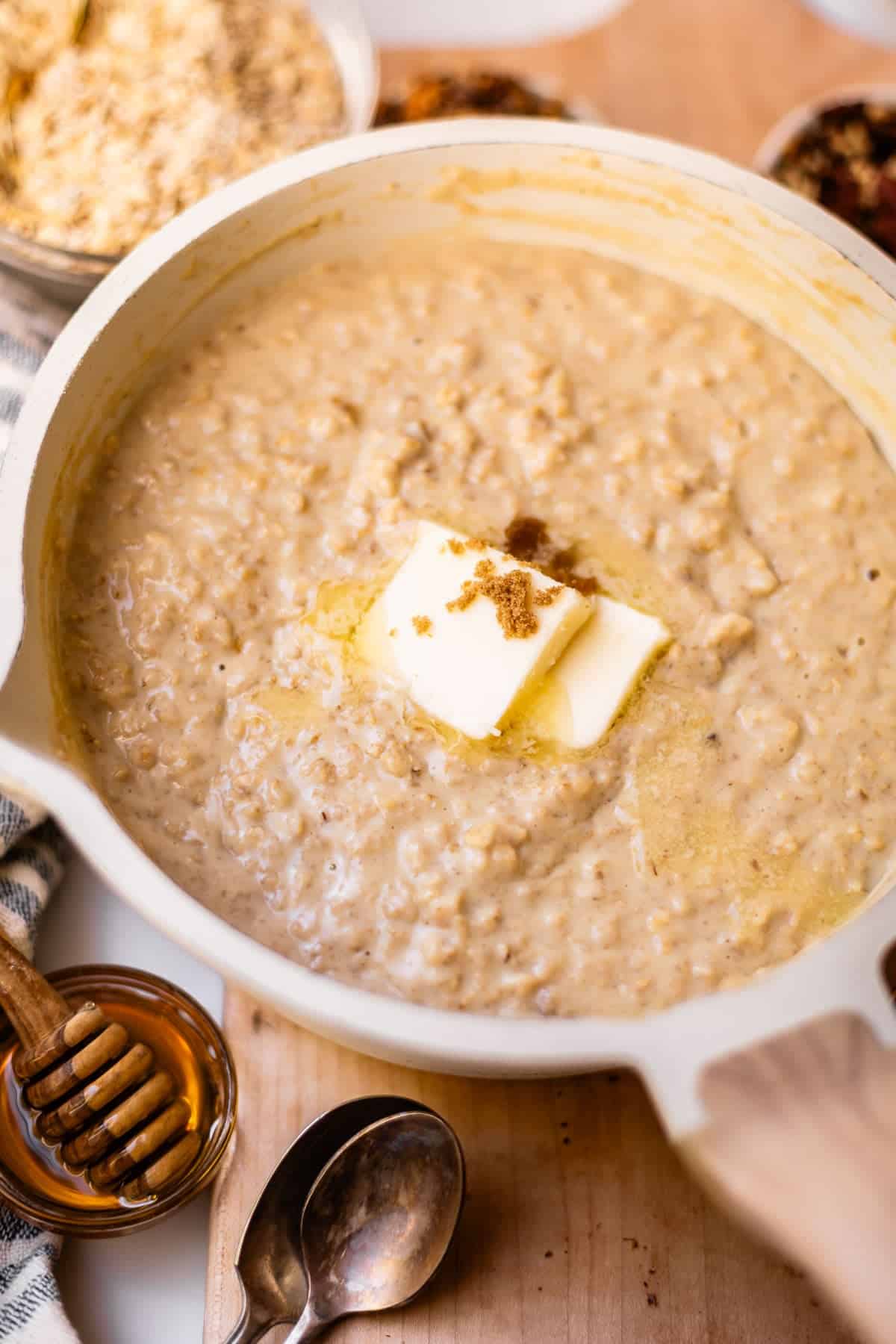 A pot of creamy oatmeal topped with two pats of butter and a sprinkle of brown sugar sits on a wooden surface, with a honey dipper, spoons, and bowls of oats and toppings nearby.