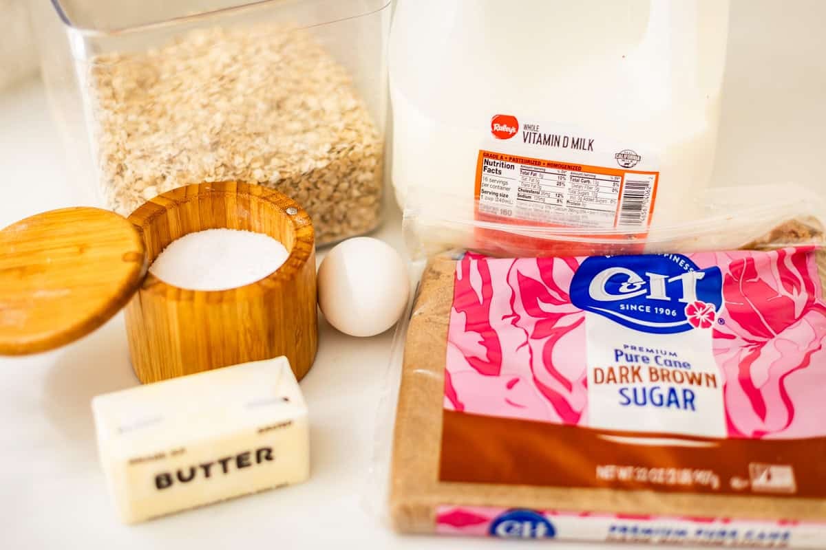 Oats, salt in a wooden container, an egg, milk, dark brown sugar, and a stick of butter are arranged on a white countertop.