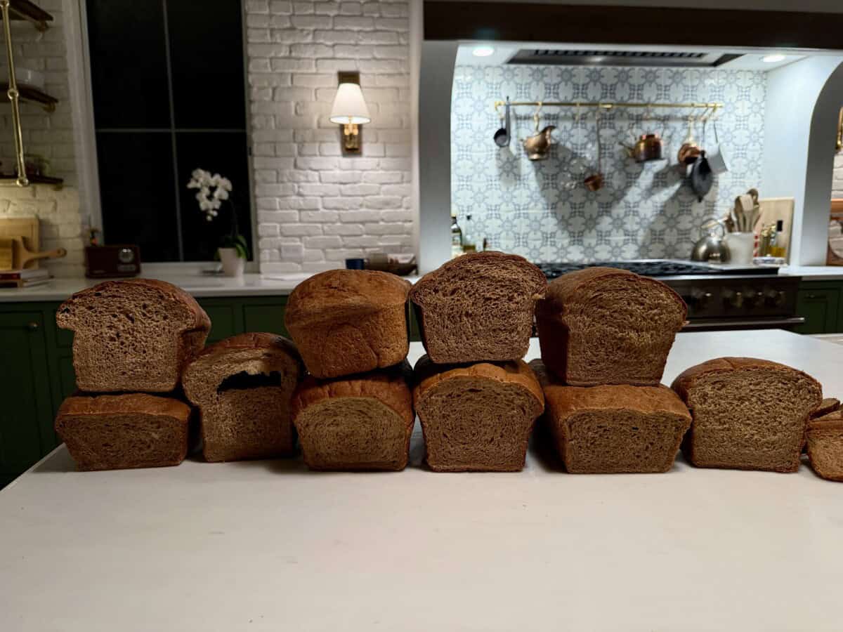 Ten loaves of wheat bread are lined up on a kitchen counter, with a modern kitchen featuring white and green cabinets, a tiled backsplash, and hanging pots in the background.