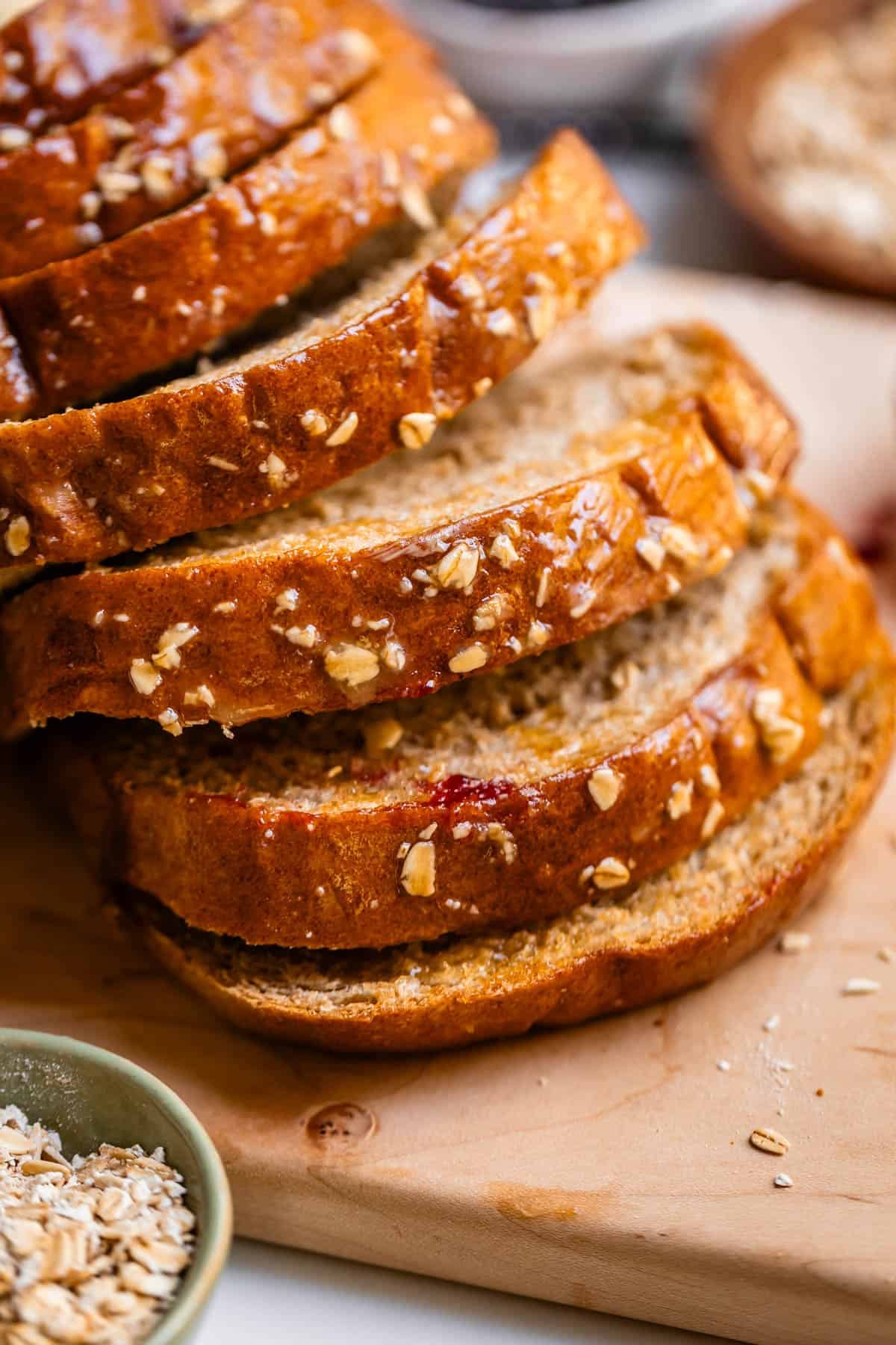 Several slices of oat-topped bread are stacked on a wooden cutting board, with a small bowl of oats beside them. The bread looks freshly baked and golden brown.